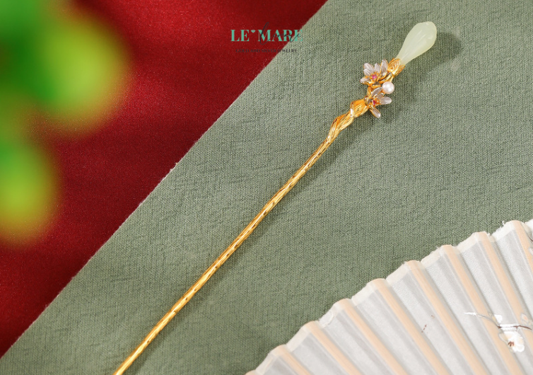 Trâm Cài Tóc Thanh Hoa – Khi nụ hoa ngọc bích chớm nở giữa mái tóc người thiếu nữ BNTC01 Thanh Hoa – cái tên gợi nhớ đến một nụ hoa vừa hé, còn giữ nguyên hơi thở trong trẻo của buổi sớm tinh khôi. Mỗi đường nét trên chiếc trâm cài tóc này đều được chạm khắc tỉ mỉ, từ cánh hoa ôm tròn như nụ cười dịu nhẹ, đến sắc ngọc bích được tuyển chọn kỹ lưỡng – xanh thẳm mà không lạnh, tươi mát mà không phai.Ngọc bích từ lâu đã được xem là viên đá quý mang năng lượng bình an và thu hút may mắn. Khi kết hợp cùng hình ảnh nụ hoa đang chúm nở, chiếc trâm cài nữ này không chỉ là điểm nhấn cho mái tóc, mà còn là biểu tượng của một tâm hồn đang độ thăng hoa – thuần khiết, dịu dàng và đầy sức sống.